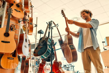 Foto de Handsome bearded curly man choosing a guitar in a musical instrument store. Hobbies and recreation. - Imagen libre de derechos