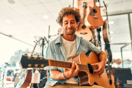 Foto de Un guapo barbudo rizado elige una guitarra en una tienda de instrumentos musicales e intenta tocarla. Pasatiempos y recreación. Comprar una guitarra en una tienda. - Imagen libre de derechos