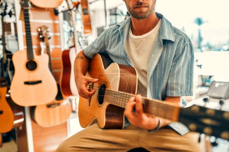 Foto de A handsome bearded curly man chooses a guitar in a musical instrument store and trying to play it. Hobbies and recreation. Buying a guitar in a store. - Imagen libre de derechos