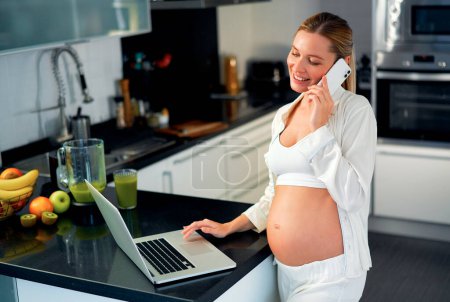 Foto de Una joven embarazada en la cocina trabajando en su portátil y hablando por teléfono, en la mesa de un batido de vitaminas recién preparado. Trabajo y alimentación saludable durante el embarazo. - Imagen libre de derechos