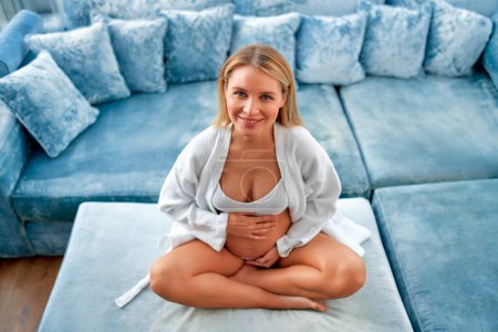 Téléchargez les photos : Une jeune femme enceinte vêtue d'un manteau blanc, les mains sur le ventre, assise sur un canapé bleu velouté près de la fenêtre de sa maison. Le concept de maternité et de grossesse. - en image libre de droit