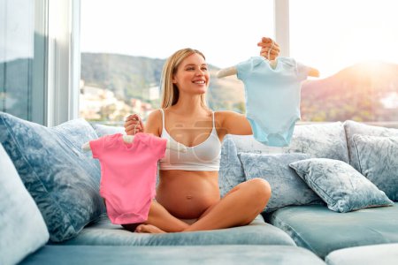 Photo for A beautiful young pregnant woman holding pink and blue baby clothes, guessing who the boy or girl will born, is sitting on the couch at home. The concept of motherhood and pregnancy. - Royalty Free Image