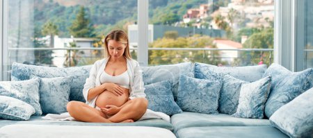 Photo for A young pregnant woman in a white coat with her hands on her tummy sitting on a velvety blue sofa by the window of her home. The concept of motherhood and pregnancy. - Royalty Free Image