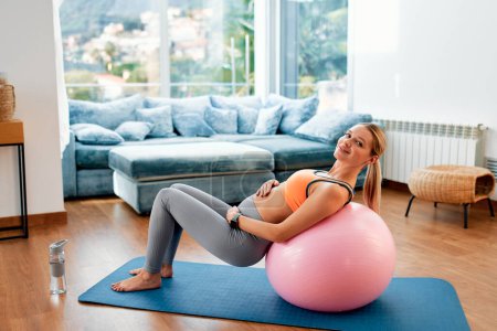 Photo for A young pregnant woman in sportswear doing exercises while sitting on a fitness ball and a rubber mat in the living room of the house. Sports during pregnancy. - Royalty Free Image