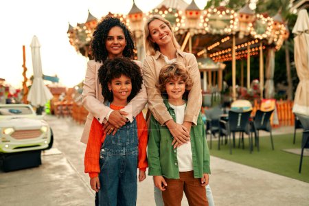 Photo for Two children with their mothers came to have fun in the amusement park. An African-American girl and a blond boy with curls are playing and have come to the park to ride a merry-go-round. - Royalty Free Image