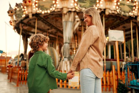 Téléchargez les photos : Un beau garçon blond enfant avec une coiffure bouclée avec sa mère est venu au parc d'attractions pour s'amuser et monter sur les carrousels et les balançoires. - en image libre de droit