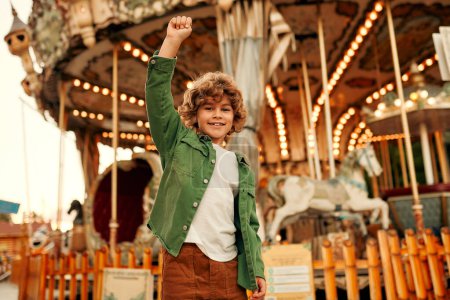 Photo for A cute Caucasian boy, a child with curly hair, came to the amusement park to have fun, play and ride a merry-go-round. A child standing in front of a merry-go-round with horses. - Royalty Free Image