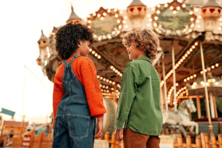 Foto de Una niña afroamericana y un niño caucásico con un peinado rizado se divierten en un parque de atracciones. - Imagen libre de derechos