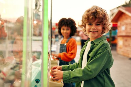 Téléchargez les photos : Les enfants jouent dans le centre d'arcade de la machine pour obtenir les griffes des jouets mous pour les pièces de monnaie. Une fille afro-américaine et un mignon garçon caucasien bouclé au parc d'attractions. - en image libre de droit