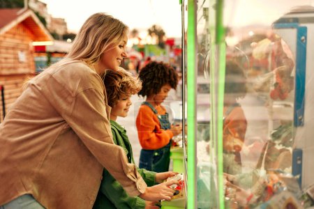 Foto de Los niños con su madre juegan en el centro de arcade de la máquina para obtener las garras de los juguetes blandos para monedas. Una chica afroamericana y un lindo chico caucásico rizado en el parque de atracciones. - Imagen libre de derechos