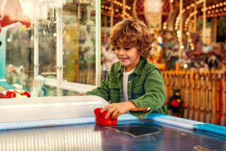 Photo for A cute Caucasian boy with curly hair playing with a puck in an air hockey in an amusement park and carousel on a weekend. - Royalty Free Image