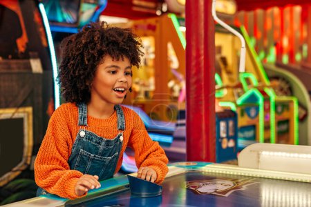 Photo for A cute African-American child with afro curls playing air hockey at an amusement park and carousel on her day off in the evening. - Royalty Free Image
