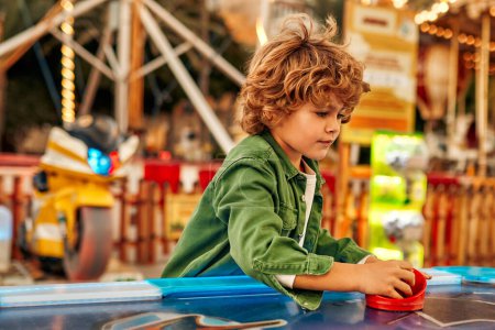 Photo for A cute Caucasian boy with curly hair playing with a puck in an air hockey in an amusement park and carousel on a weekend. - Royalty Free Image