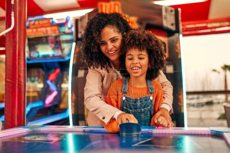 Photo for A cute African-American child with afro curls with her mother playing air hockey at an amusement park and carousel on her day off in the evening. - Royalty Free Image