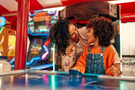Photo for A cute African-American child with afro curls with her mother playing air hockey at an amusement park and carousel on her day off in the evening. - Royalty Free Image