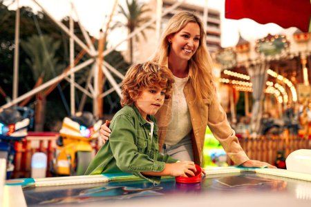 Photo for A cute Caucasian boy with curly hair with his mother playing with a puck in an air hockey in an amusement park and carousel on a weekend. - Royalty Free Image