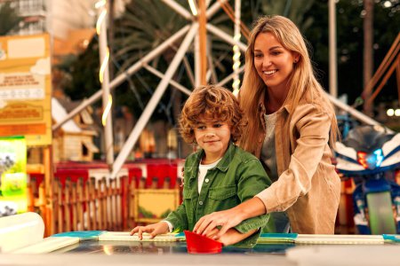 Photo for A cute Caucasian boy with curly hair with his mother playing with a puck in an air hockey in an amusement park and carousel on a weekend. - Royalty Free Image