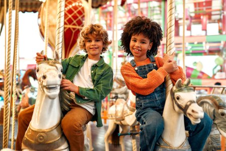 Photo for An African-American girl with Afro-curls and a Caucasian curly-haired blonde boy ride a vintage horse carousel in the evening at an amusement park on a weekend. - Royalty Free Image