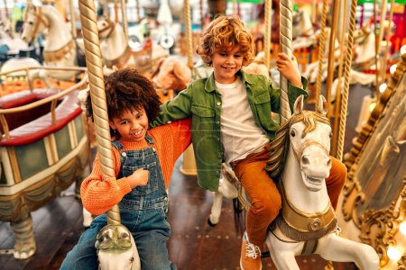 Téléchargez les photos : Une fille afro-américaine avec des afro-boucles et un garçon blond aux cheveux bouclés caucasiens monte un carrousel de cheval vintage le soir dans un parc d'attractions un week-end. - en image libre de droit
