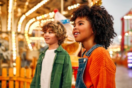 Téléchargez les photos : Une fille afro-américaine avec des boucles afro, et un garçon caucasien regardent admirablement un carrousel de chevaux dans un parc d'attractions ou un cirque un week-end soir. - en image libre de droit