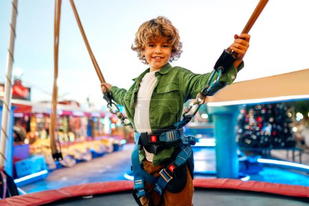 Foto de Un niño caucásico saltando sobre un trampolín con bandas elásticas de seguros en un parque de diversiones y carruseles. - Imagen libre de derechos
