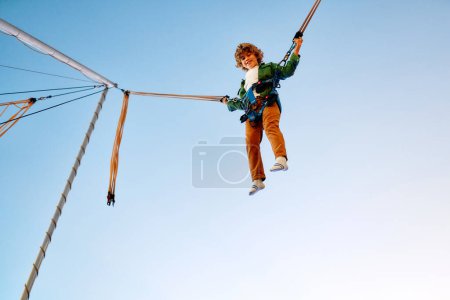 Photo for A Caucasian boy child jumping on a trampoline with insurance elastic bands in an amusement park and carousels. - Royalty Free Image