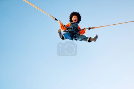 Photo for An african american girl child with afro curly hair jumping on a trampoline with insurance elastic bands in an amusement park and carousels. - Royalty Free Image