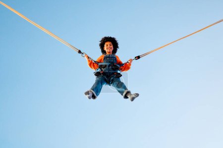Photo for An african american girl child with afro curly hair jumping on a trampoline with insurance elastic bands in an amusement park and carousels. - Royalty Free Image
