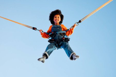 Foto de Una niña afroamericana con el pelo rizado afro saltando en un trampolín con bandas elásticas de seguros en un parque de atracciones y carruseles. - Imagen libre de derechos