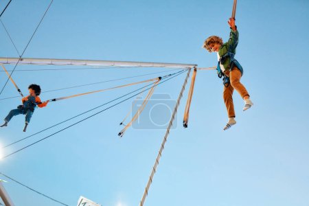 Photo for An african american girl child with afro curly hair and caucasian cute boy jumping on a trampoline with insurance elastic bands in an amusement park and carousels. - Royalty Free Image