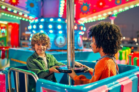 Photo for An African-American sweet girl child with afro curls and a cute Caucasian boy ride together on a carousel in an amusement park in the evening on weekend and have fun. - Royalty Free Image