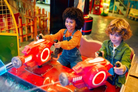 Photo for An African-American girl child with an Afro hairstyle and a cute Caucasian boy sitting in an amusement car ride playing a computer game racing in an amusement park in the evening laughing merrily and relaxing together. - Royalty Free Image