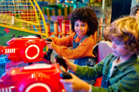 Téléchargez les photos : Une fille afro-américaine enfant avec une coiffure afro et un mignon garçon caucasien assis dans une promenade en voiture de divertissement jouer à un jeu d'ordinateur de course dans un parc d'attractions le soir en riant joyeusement et en se relaxant ensemble. - en image libre de droit