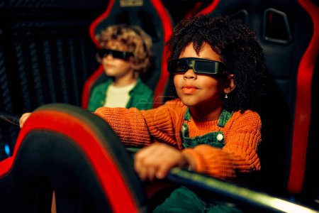 Foto de Una chica afroamericana y un lindo chico caucásico sentados en un cine virtual en gafas en movimiento sillas giratorias y emocionalmente viendo una película en un parque de atracciones. - Imagen libre de derechos