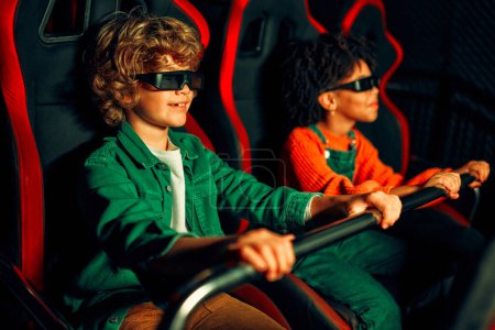 Foto de Una chica afroamericana y un lindo chico caucásico sentados en un cine virtual en gafas en movimiento sillas giratorias y emocionalmente viendo una película en un parque de atracciones. - Imagen libre de derechos