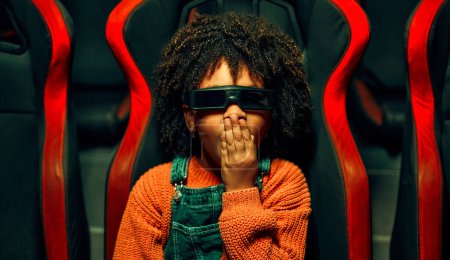 Foto de Una chica linda afroamericana con y un chico caucásico lindo sentado en un cine virtual en gafas en movimiento sillas giratorias y emocionalmente viendo una película en un parque de atracciones. - Imagen libre de derechos