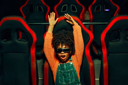 Foto de Una chica linda afroamericana con y un chico caucásico lindo sentado en un cine virtual en gafas en movimiento sillas giratorias y emocionalmente viendo una película en un parque de atracciones. - Imagen libre de derechos