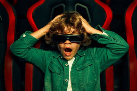 Foto de Un lindo chico caucásico con el pelo rizado sentado en un cine virtual en gafas en movimiento sillas giratorias y emocionalmente viendo una película en un parque de atracciones. - Imagen libre de derechos