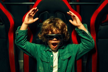Téléchargez les photos : Un mignon garçon caucasien aux cheveux bouclés assis dans un cinéma virtuel à lunettes sur des chaises tournantes mobiles et regardant émotionnellement un film dans un parc d'attractions. - en image libre de droit
