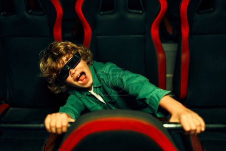 Téléchargez les photos : Un mignon garçon caucasien aux cheveux bouclés assis dans un cinéma virtuel à lunettes sur des chaises tournantes mobiles et regardant émotionnellement un film dans un parc d'attractions. - en image libre de droit
