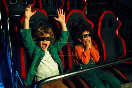 Téléchargez les photos : Une fille afro-américaine et un mignon garçon caucasien assis dans un cinéma virtuel à lunettes sur des chaises tournantes mobiles et regardant émotionnellement un film dans un parc d'attractions. - en image libre de droit
