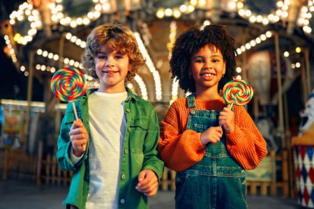 Photo for A cute African-American girl with an Afro hairstyle and caucasian boy eating a colorful lollipop standing against the background of a carousel with horses in the evening at an amusement park or circus. - Royalty Free Image