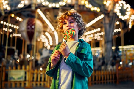 Téléchargez les photos : Un mignon garçon caucasien aux cheveux bouclés blonds mangeant une sucette colorée debout sur le fond d'un carrousel avec des chevaux le soir dans un parc d'attractions ou un cirque. - en image libre de droit