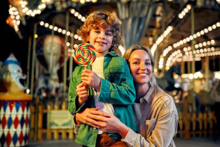 Photo for A cute caucasian boy with blonde curly hair with his mother eating a colorful lollipop standing against the background of a carousel with horses in the evening at an amusement park or circus. - Royalty Free Image