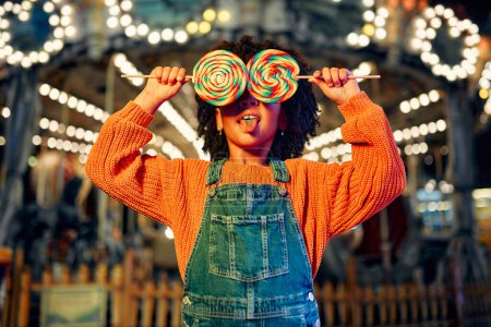 Téléchargez les photos : Une jolie fille afro-américaine avec une coiffure afro mange une sucette colorée debout sur le fond d'un carrousel avec des chevaux le soir dans un parc d'attractions ou un cirque. - en image libre de droit