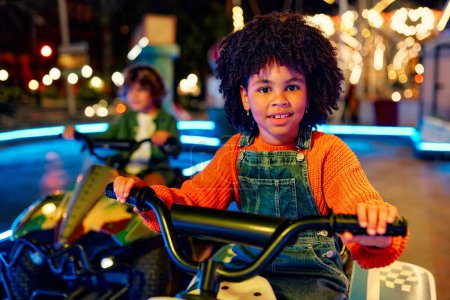 Foto de Una chica afroamericana y un lindo chico caucásico montando un coche juntos en la noche en un carrusel y parque de atracciones. - Imagen libre de derechos