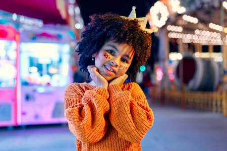 Téléchargez les photos : Une jolie fille afro-américaine avec une coiffure afro, des paillettes sur les joues et une couronne sur la tête comme une princesse debout devant des carrousels rougeoyants dans un parc d'attractions le soir. - en image libre de droit
