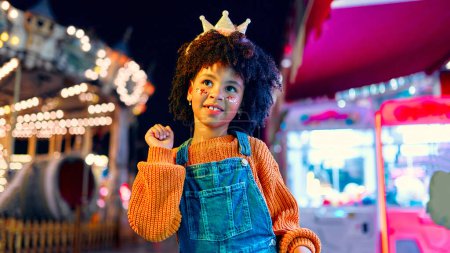 Foto de Una linda chica afroamericana con un peinado afro, lentejuelas en sus mejillas y una corona en su cabeza como una princesa de pie frente a brillantes carruseles en un parque de atracciones por la noche. - Imagen libre de derechos