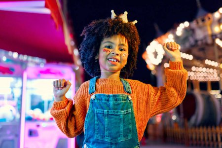 Photo for A cute African-American girl with an Afro hairstyle, sequins on her cheeks and a crown on her head like a princess standing in front of glowing carousels at an amusement park in the evening. - Royalty Free Image