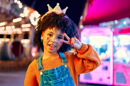 Téléchargez les photos : Une jolie fille afro-américaine avec une coiffure afro, des paillettes sur les joues et une couronne sur la tête comme une princesse debout devant des carrousels rougeoyants dans un parc d'attractions le soir. - en image libre de droit